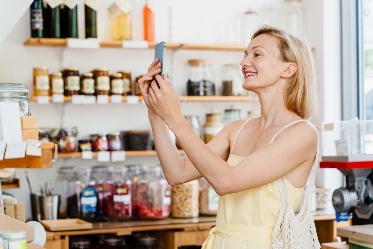 Femeie care fotografiază cu telefonul rafturile unui magazin local | GetVizual